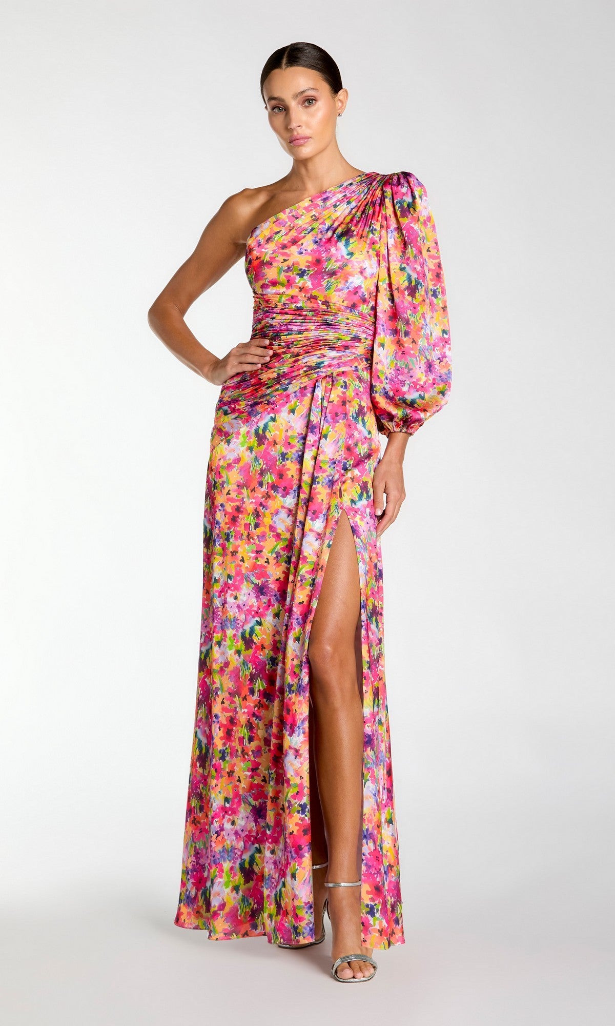 A woman stands elegantly against a plain background, wearing the Mac Duggal 2634 One-Sleeve Long Print Formal Dress with a ruched bodice, vibrant floral print, puff sleeve, and thigh-high slit.